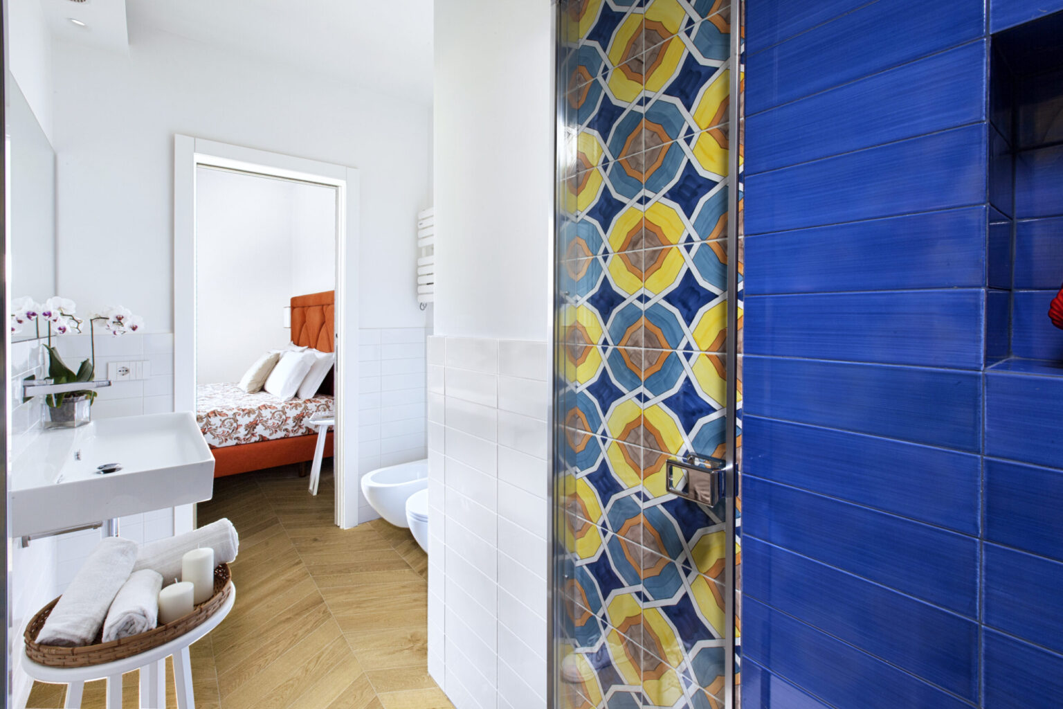 Modern bathroom interior featuring a vibrant blue tiled shower wall with yellow and orange geometric patterns, a sleek white sink with towels, and a cozy bedroom visible in the background. | Only on SopranoVillas