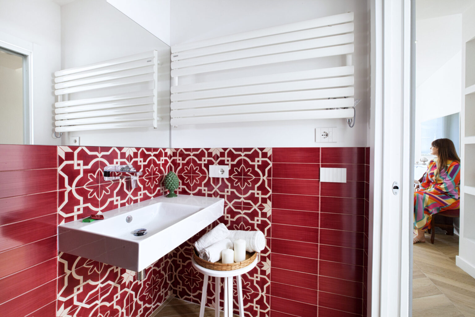 Modern bathroom with red patterned tiles, a sleek white sink, and stylish towel radiator, featuring a woman in a colorful robe in the background. | Only on SopranoVillas