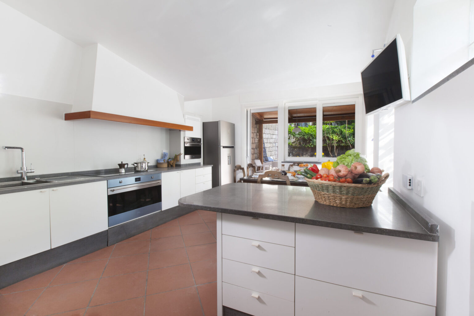 Modern kitchen with sleek white cabinetry, stainless steel appliances, and a spacious countertop featuring a basket of fresh fruits, illuminated by natural light from large windows. | Only on SopranoVillas