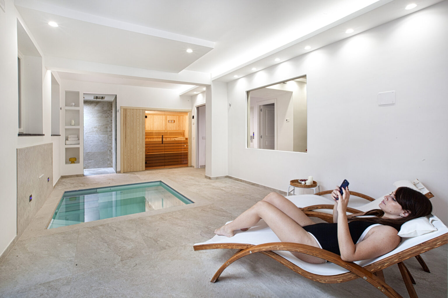 Relaxing woman in a spa-like room with a small indoor pool, lounging on a wooden chair while using a smartphone, showcasing a modern and tranquil wellness environment. | Only on SopranoVillas