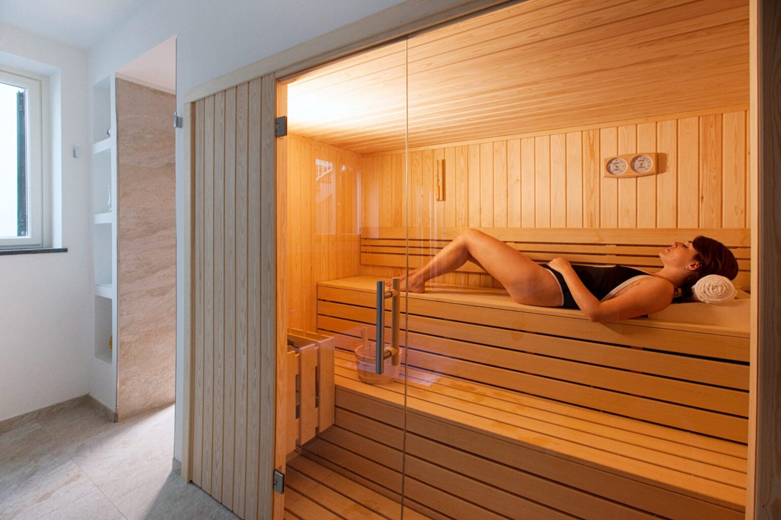 Woman relaxing in a wooden sauna, enjoying the warm ambiance and therapeutic benefits of heat therapy. The sauna features wooden benches and soft lighting, creating a tranquil environment for wellness and relaxation. | Only on SopranoVillas