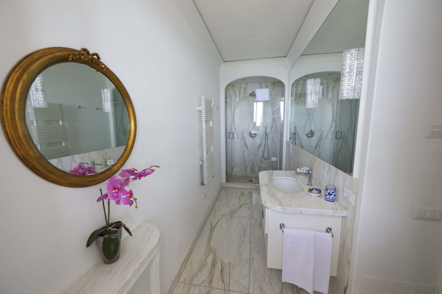 Modern bathroom interior featuring a marble floor, a stylish sink, and a round decorative mirror. The space is bright and airy, with glass shower enclosures and a touch of elegance from a purple orchid arrangement. | Only on SopranoVillas