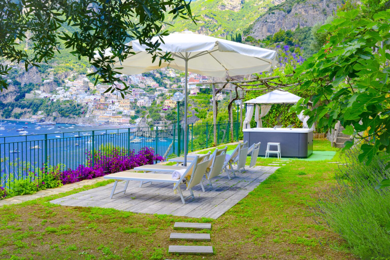 Scenic outdoor patio with lounge chairs and umbrellas overlooking a coastal view, surrounded by greenery and vibrant flowers. Ideal for relaxation and enjoying the serene landscape. | Only on SopranoVillas