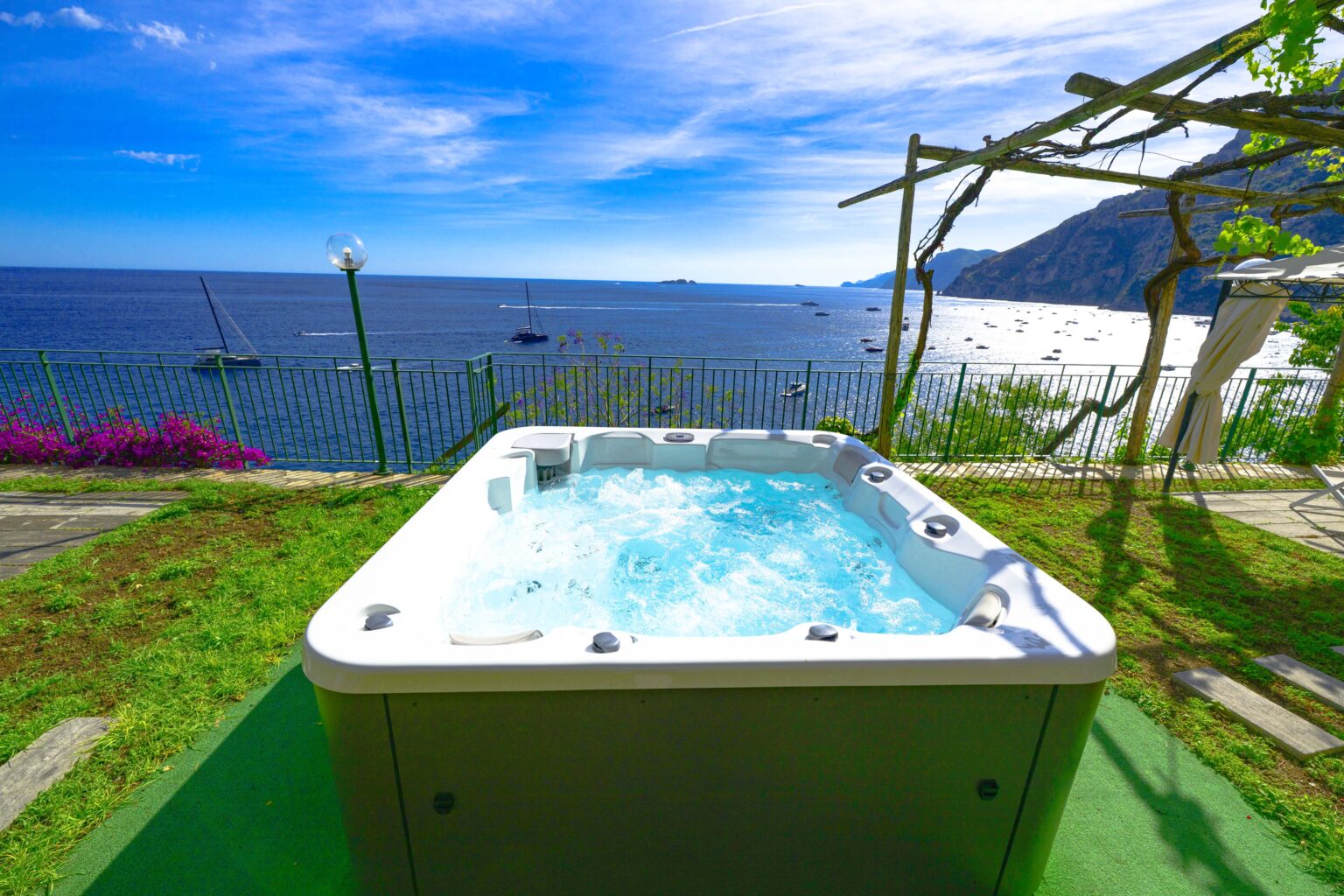 Hot tub overlooking the ocean, surrounded by lush greenery and vibrant flowers, with a clear blue sky and distant boats on the water, ideal for relaxation and luxury getaway. | Only on SopranoVillas