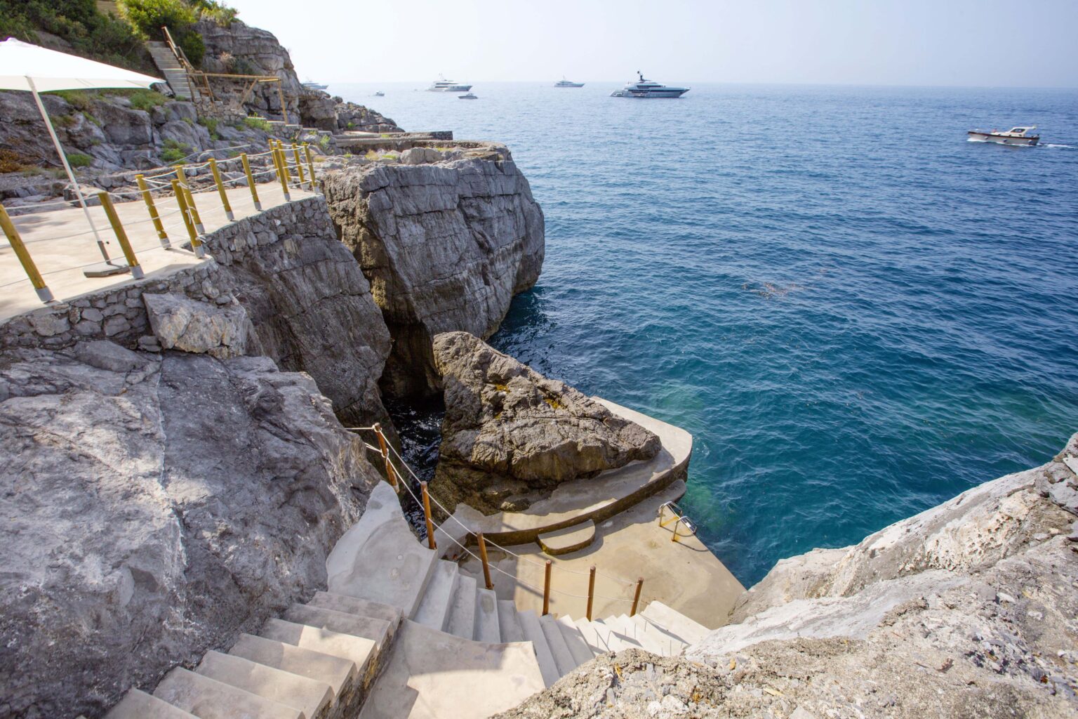 Staircase leading down to a rocky shoreline with clear blue water and boats in the distance, showcasing a scenic coastal view. | Only on SopranoVillas