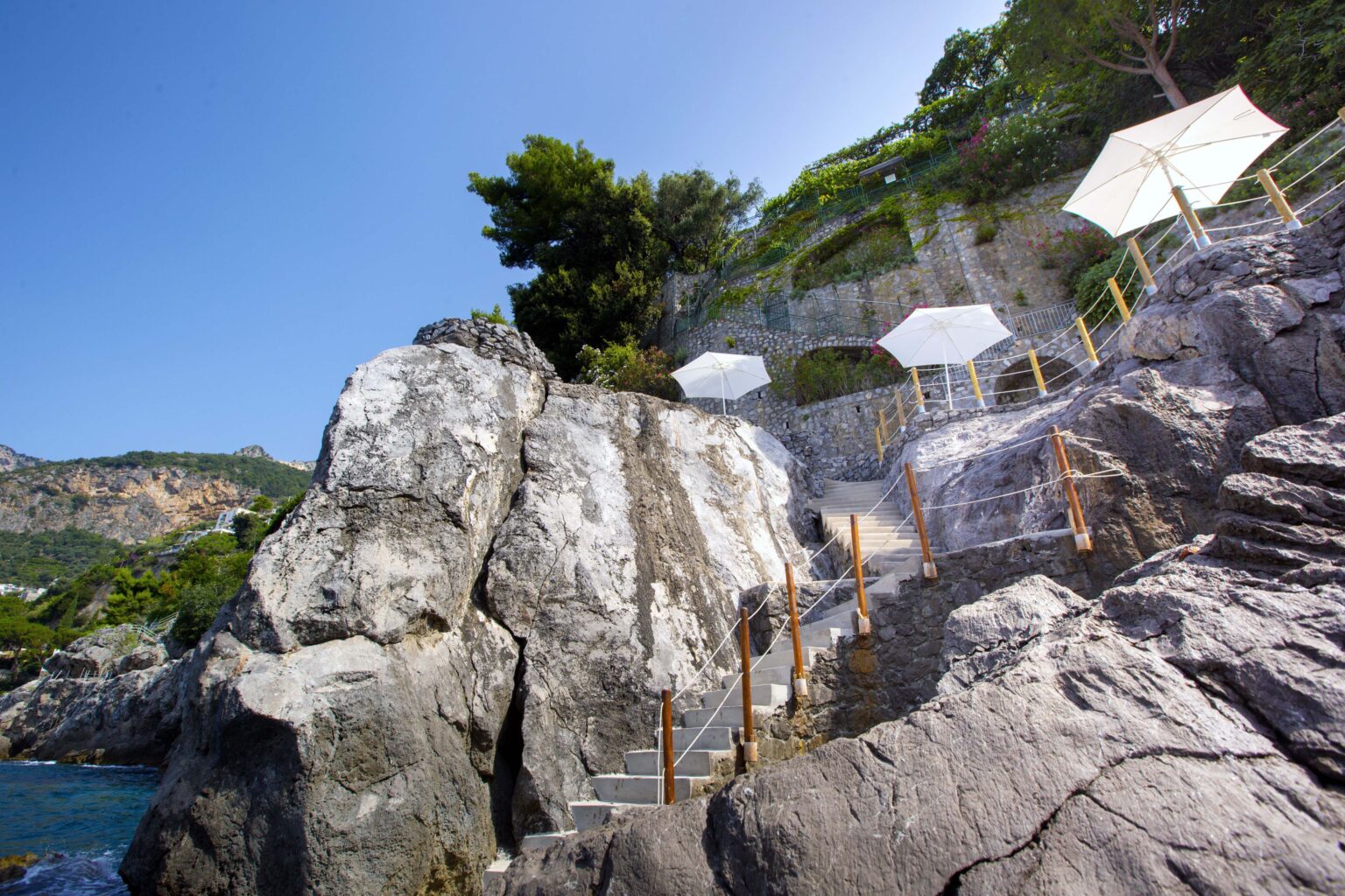 Scenic coastal view featuring rocky terrain with wooden steps leading to a serene beach area, complemented by white umbrellas for shade and lush greenery in the background. Ideal for vacation and relaxation. | Only on SopranoVillas
