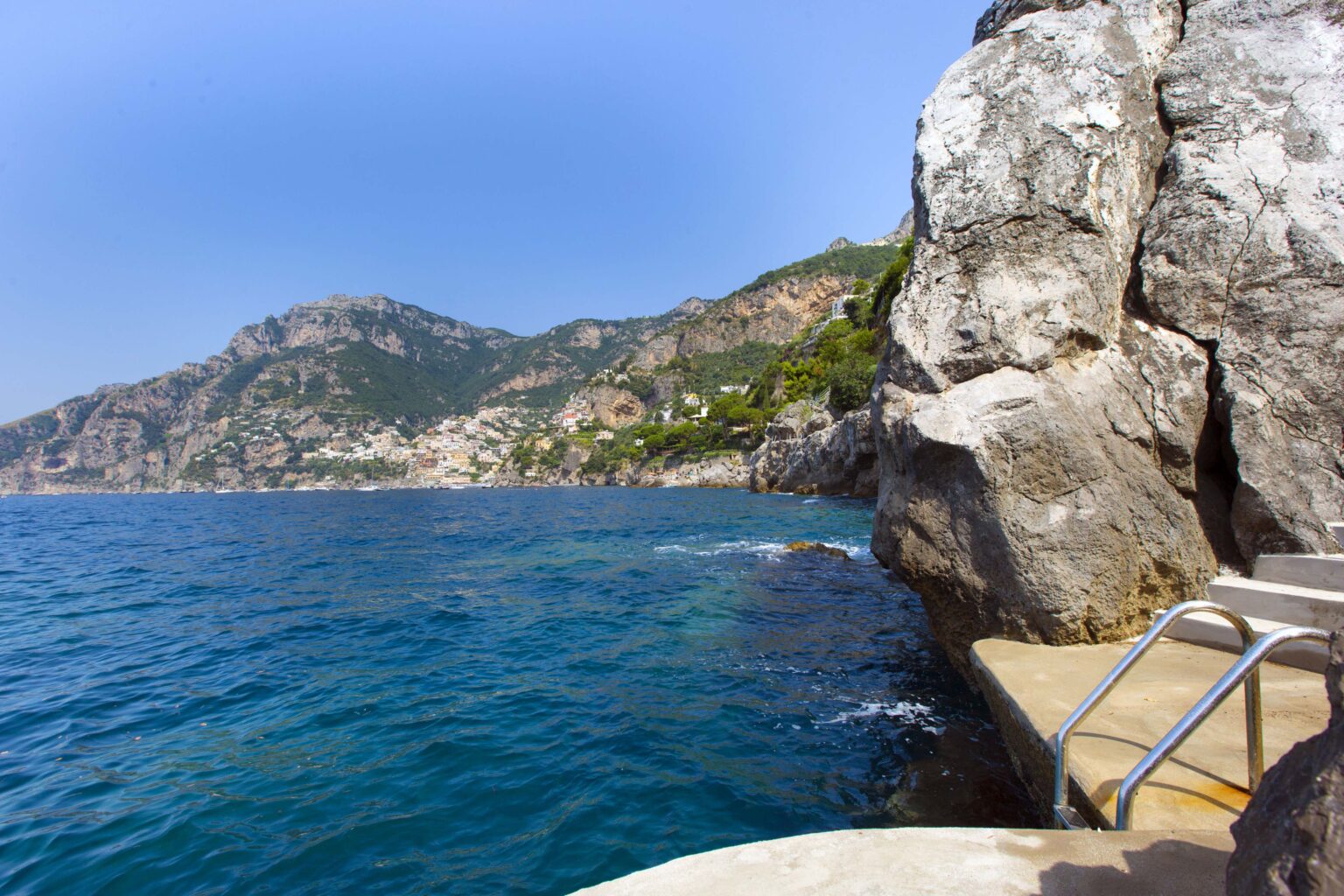 Scenic view of a rocky coastline with clear blue water, showcasing mountains and a distant village along the Amalfi Coast in Italy. | Only on SopranoVillas