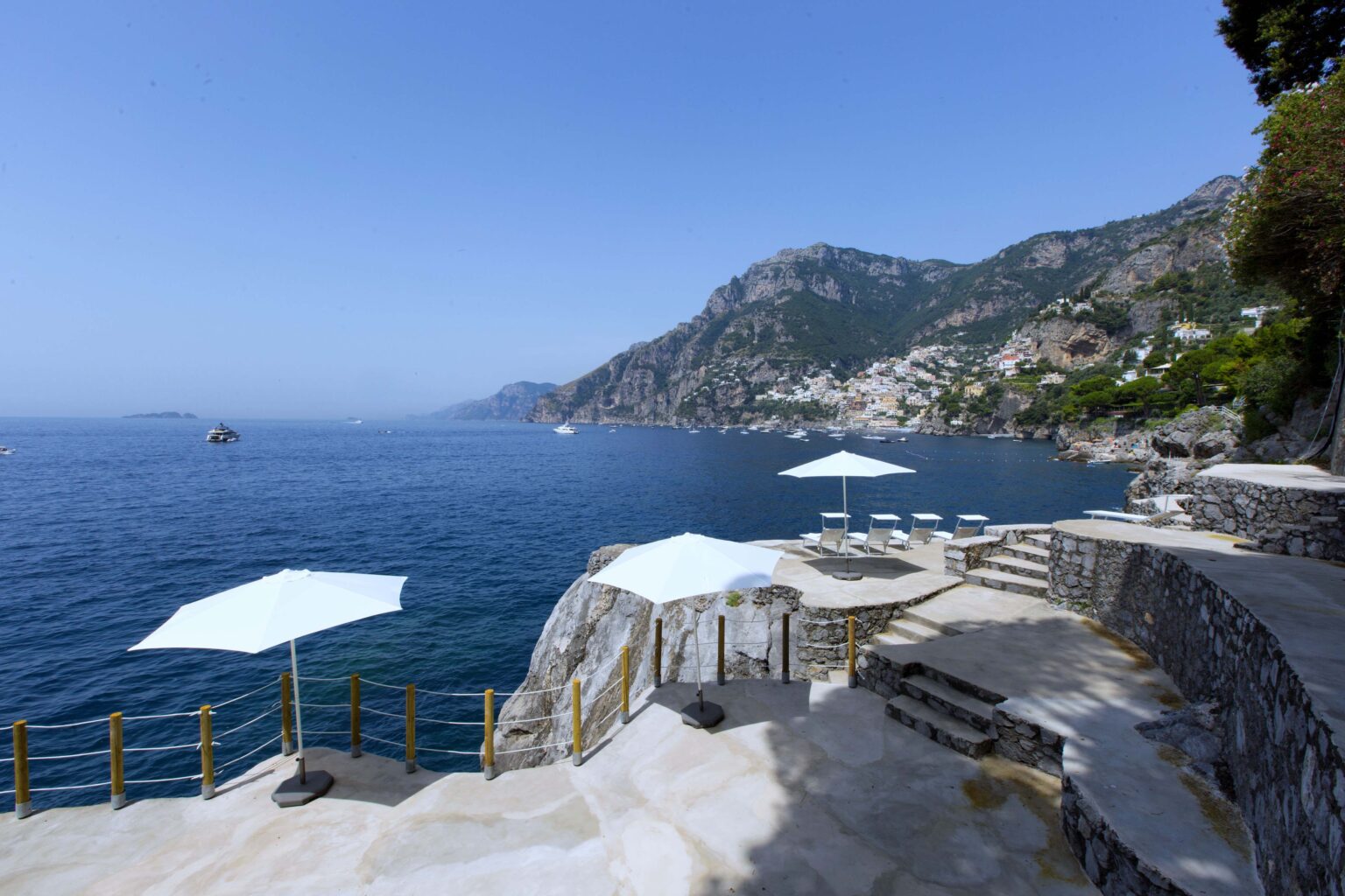Scenic view of a coastal area featuring white umbrellas and stone steps leading to the crystal-clear blue water, with rocky cliffs and a distant village in the background under a clear sky. | Only on SopranoVillas