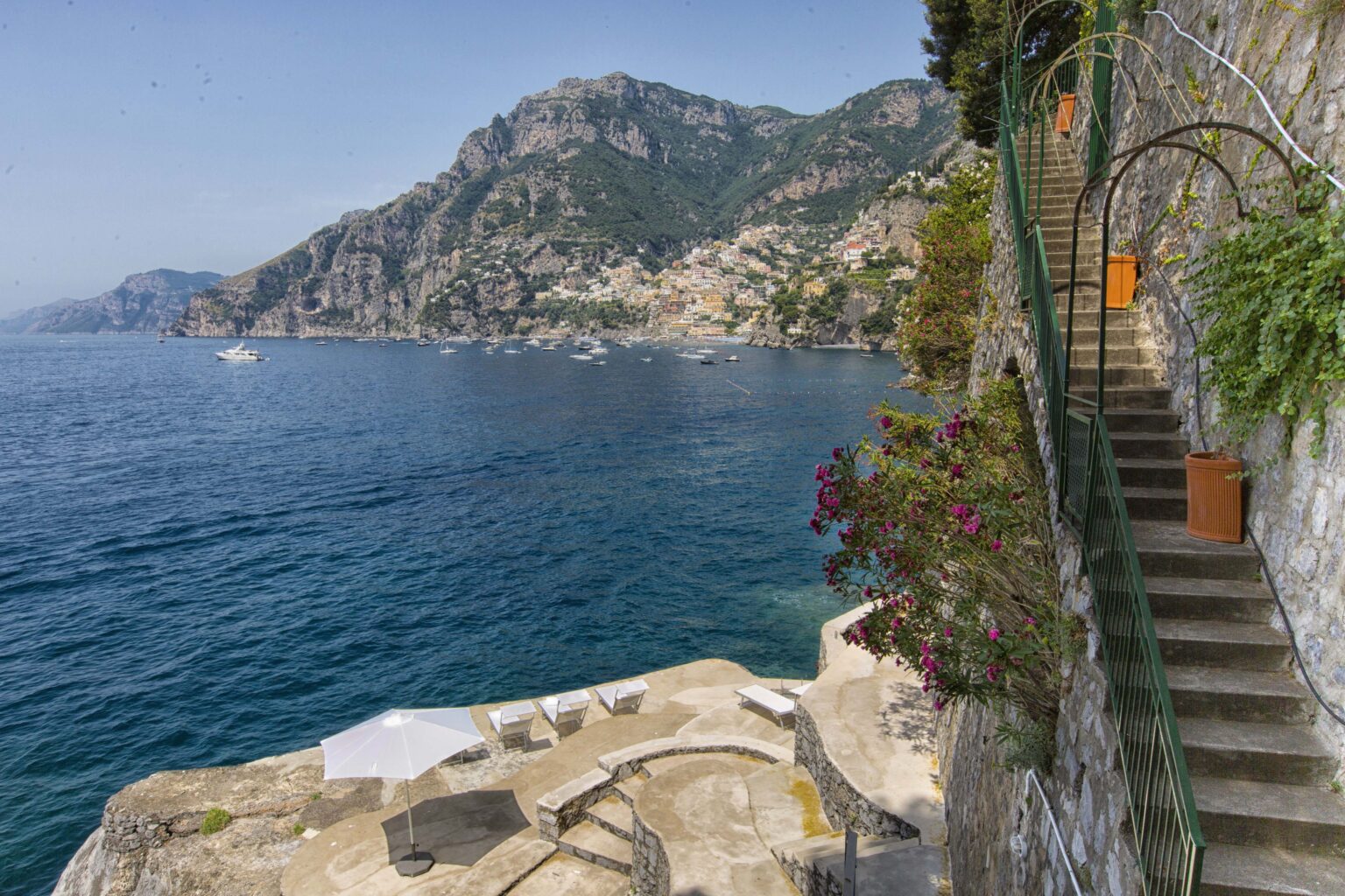 Scenic view of the Amalfi Coast featuring a stone staircase leading down to the turquoise sea, surrounded by lush greenery and colorful flowers, with rocky cliffs and distant coastal towns in the background. | Only on SopranoVillas
