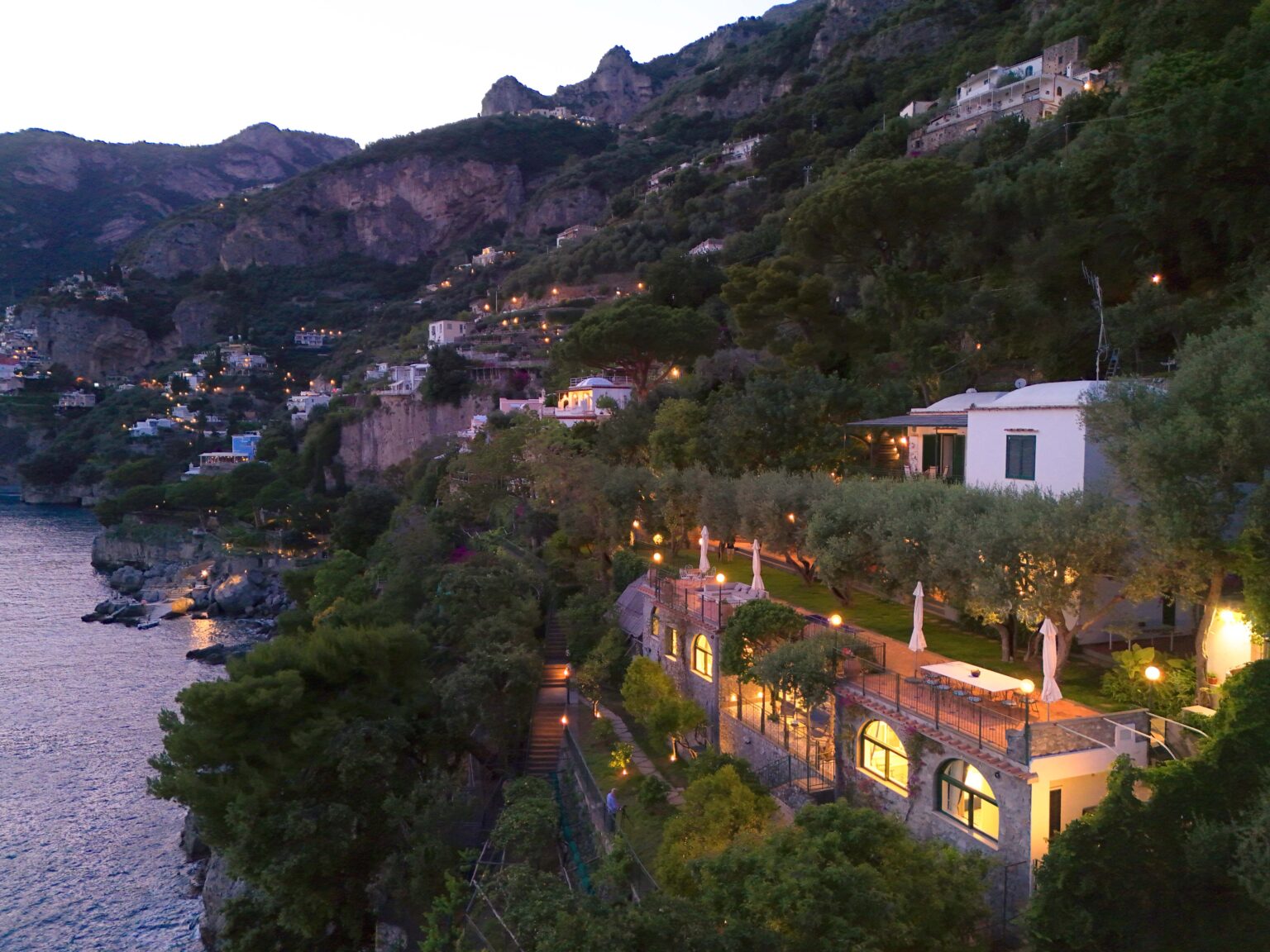Luxury villa for rent in Italy, featuring stunning coastal views, lush gardens, and elegant architecture, set against the backdrop of a picturesque hillside at dusk. Perfect for a serene getaway. | Only on SopranoVillas