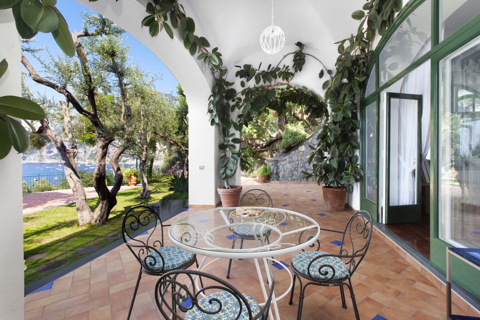 Luxury villa terrace in Italy featuring a stylish dining area surrounded by lush greenery and stunning views of the garden and coastline. Ideal for relaxing and enjoying outdoor dining in a serene setting. | Only on SopranoVillas