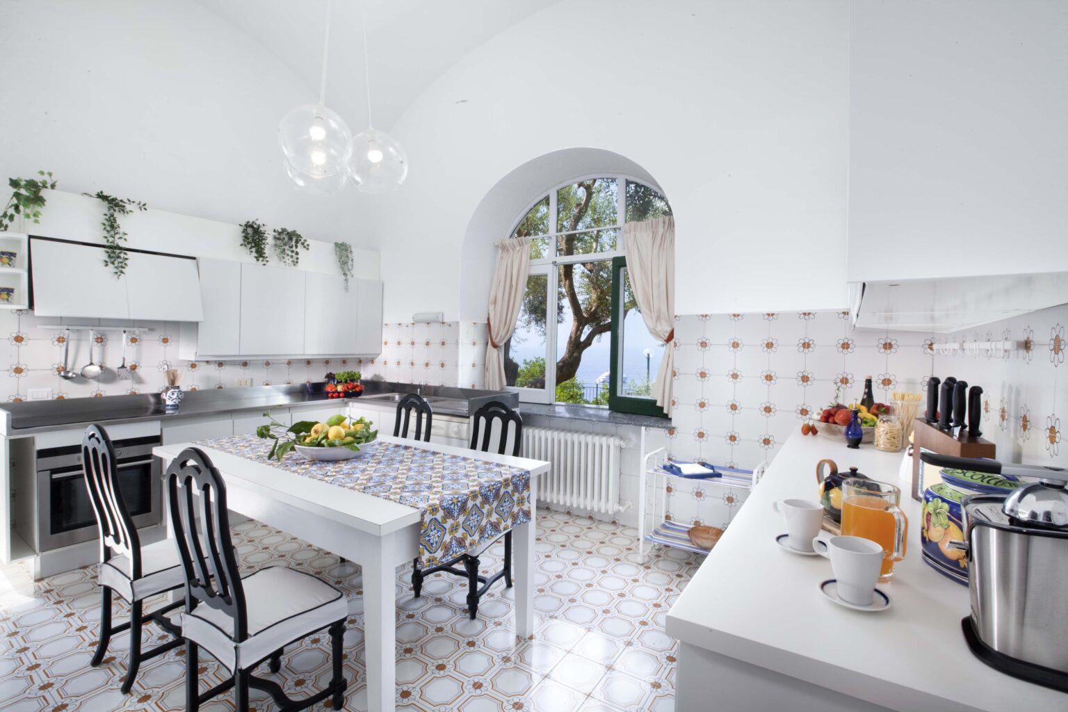 Bright and modern kitchen featuring white cabinetry, a spacious dining table with black chairs, and large windows that provide natural light and a view of greenery outside. The tiled floor adds a classic touch, while kitchen appliances and decor enhance functionality and style. | Only on SopranoVillas