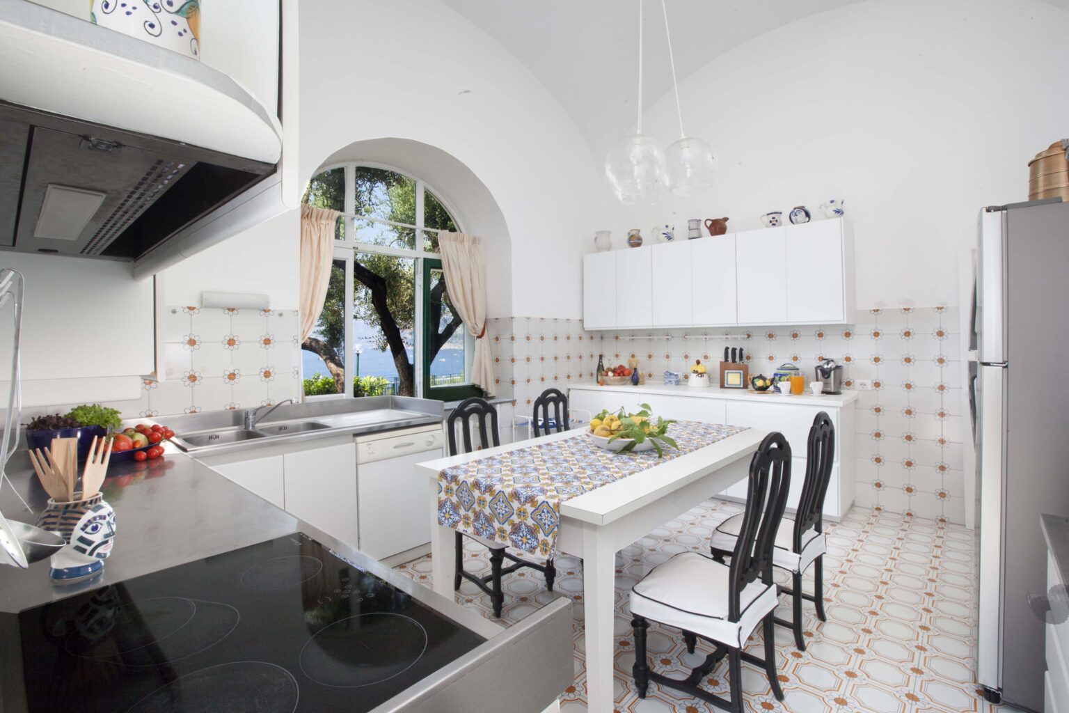 Bright and spacious modern kitchen featuring white cabinetry, a central dining table with decorative tablecloth, and large windows that provide natural light. The kitchen is equipped with stainless steel appliances and has a clean, minimalist design, ideal for cooking and entertaining. | Only on SopranoVillas