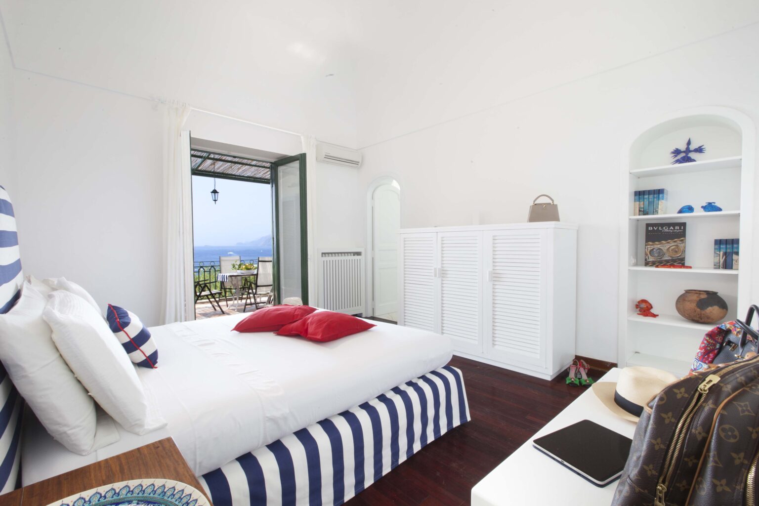 Cozy bedroom with white walls and striped blue bedding, featuring red accent pillows. The room includes a wardrobe, a bookshelf with decorative items, and a view of the outdoors through an open door. Perfect for a relaxing getaway. | Only on SopranoVillas