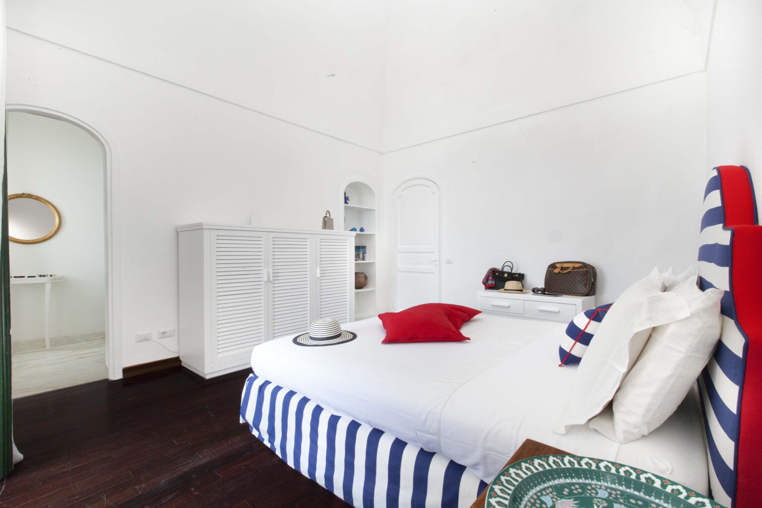 Bright and airy bedroom featuring a white color scheme, striped blue and white bedding, a red accent pillow, and modern decor. The room includes a stylish dresser and a round mirror, enhancing the overall aesthetic. Perfect for showcasing contemporary interior design. | Only on SopranoVillas