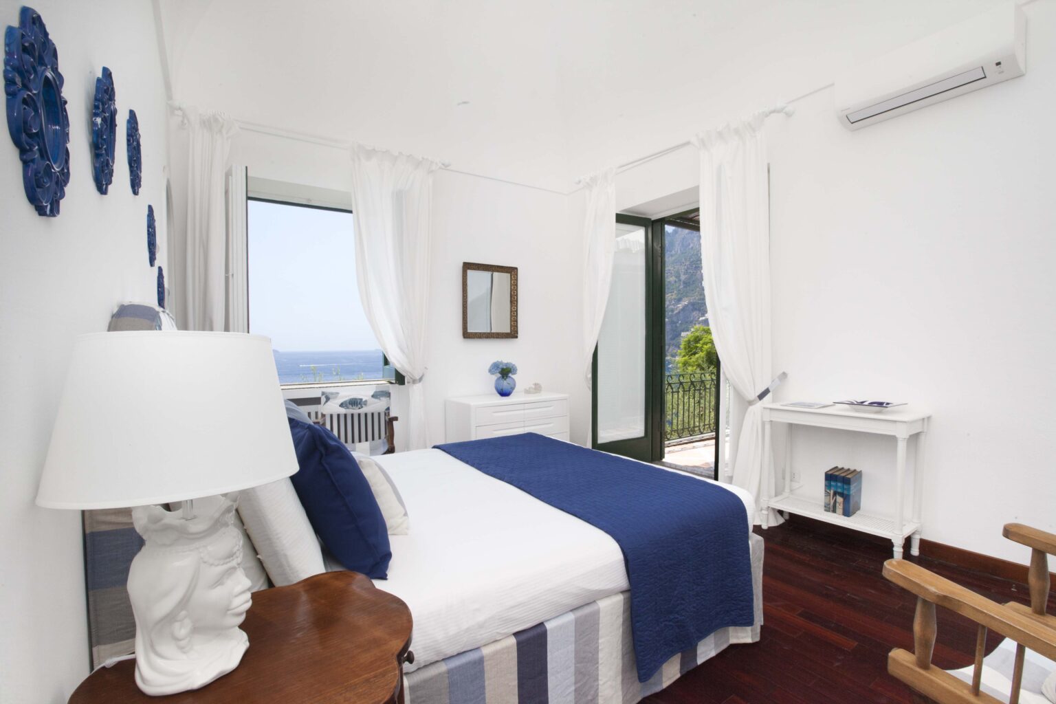Cozy bedroom with white walls and blue accents, featuring a comfortable bed, a stylish lamp, and a desk. Large windows provide a scenic view of the outdoors, enhancing the room's airy and bright atmosphere. Ideal for relaxation and comfort. | Only on SopranoVillas