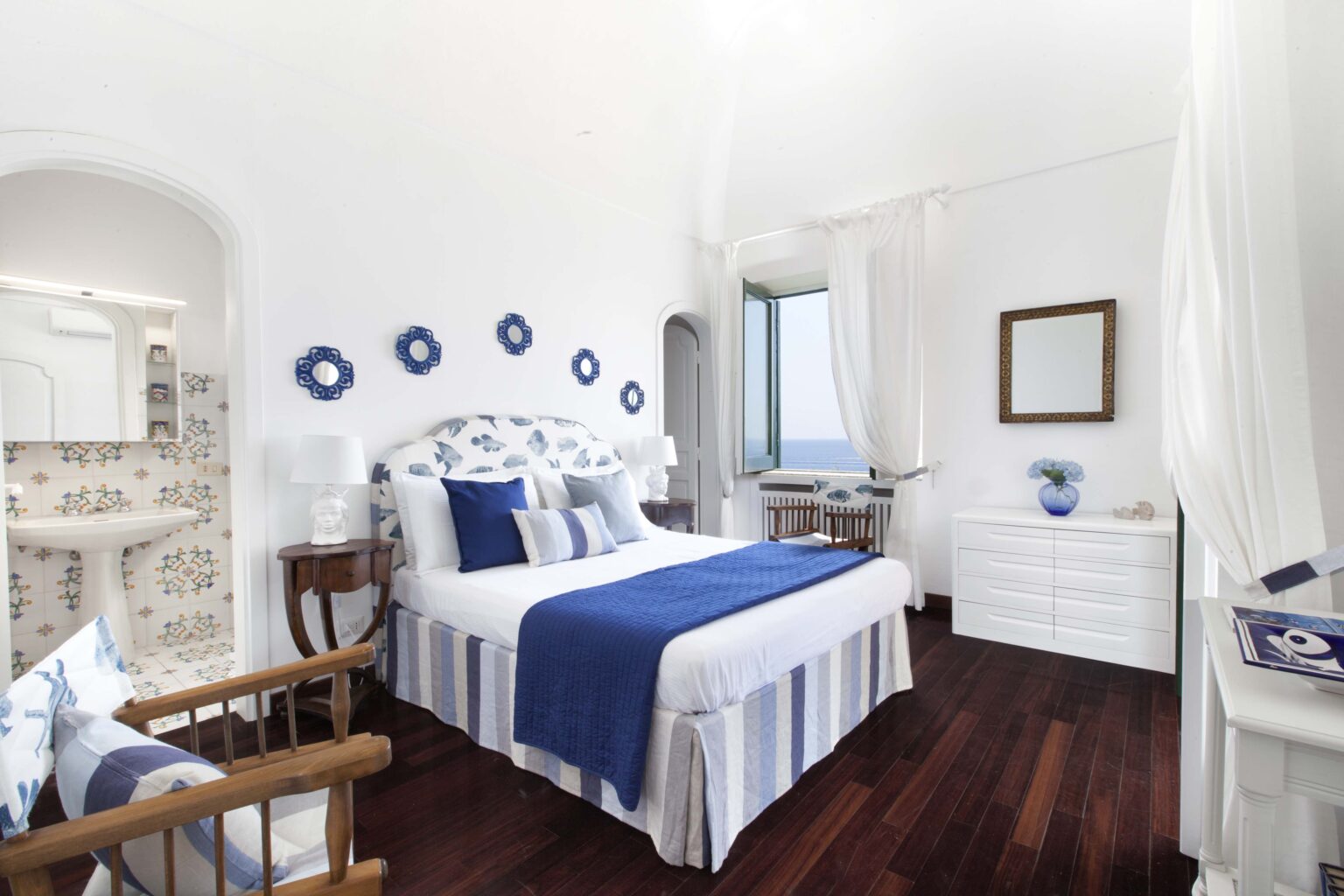 Cozy and modern bedroom featuring a white color scheme with blue accents, a comfortable bed with decorative pillows, wooden flooring, and natural light streaming through large windows. Ideal for relaxation and interior design inspiration. | Only on SopranoVillas