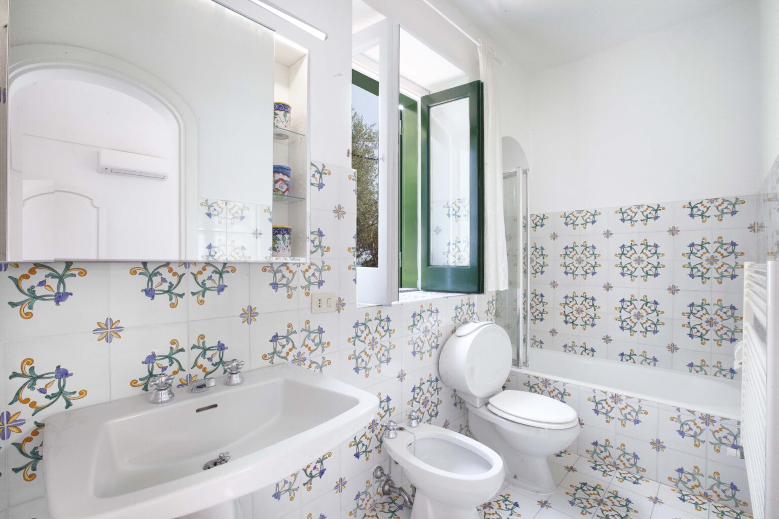 Modern bathroom featuring a stylish design with patterned ceramic tiles, a white sink, a toilet, and a bathtub. Natural light floods the space through a window, enhancing the bright and airy atmosphere. | Only on SopranoVillas