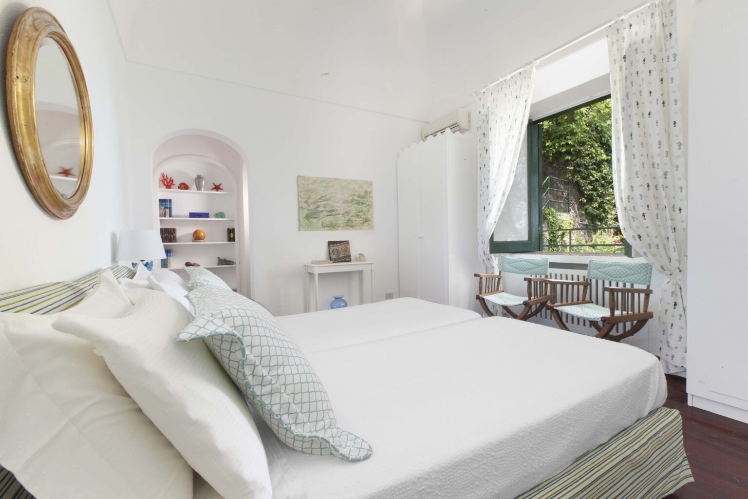 Bright and airy bedroom featuring a comfortable bed with decorative pillows, large windows with sheer curtains, and a cozy seating area. The room is decorated in light colors with a touch of greenery visible outside, creating a serene atmosphere. | Only on SopranoVillas