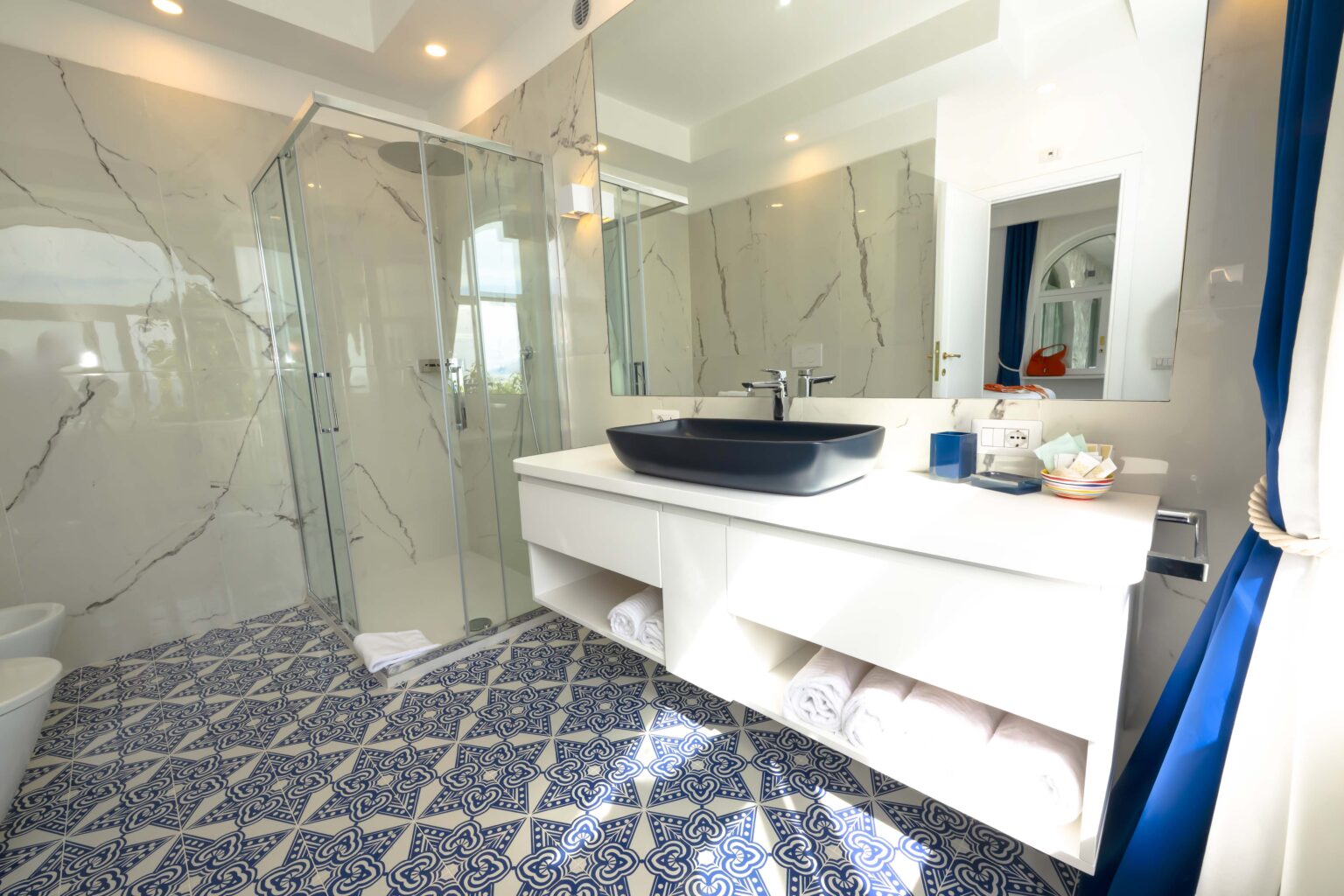 Modern luxury bathroom in an Italian villa for rent, featuring elegant marble walls, a spacious glass shower, a stylish dark basin, and intricate patterned tiles. Ideal for a sophisticated getaway. | Only on SopranoVillas