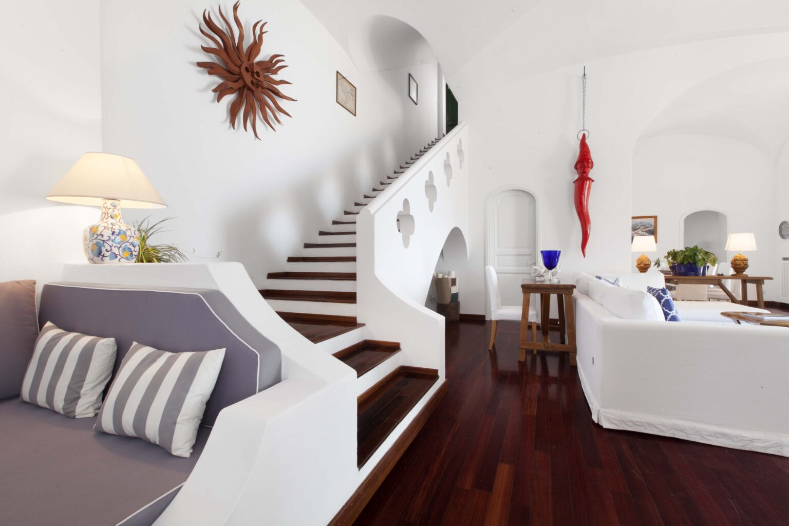 Bright and airy interior of a modern living space featuring a staircase, white walls, wooden flooring, and stylish furniture. The room is decorated with a sunburst wall art and has ample natural light, creating a welcoming atmosphere. | Only on SopranoVillas