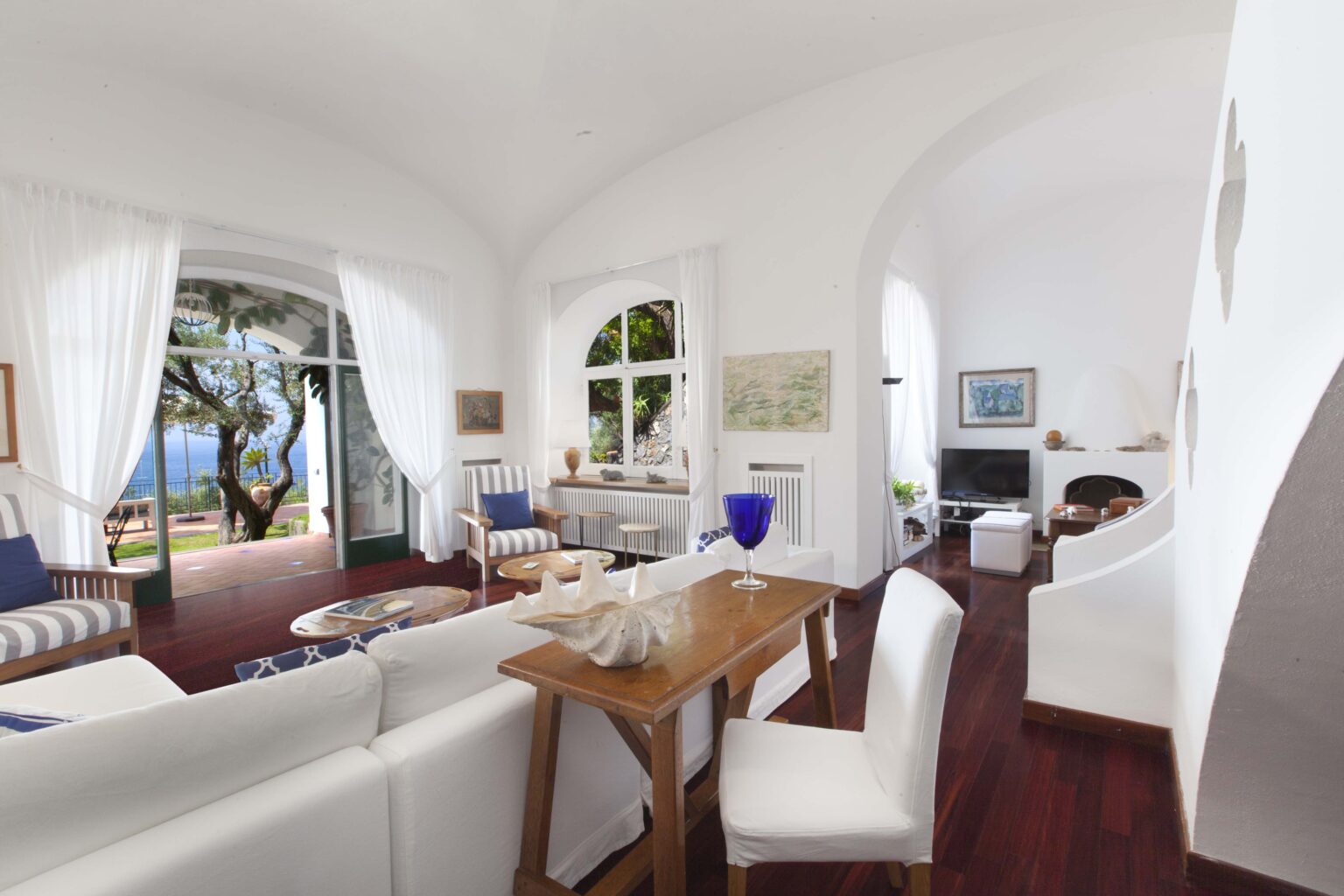 Bright and airy living room with white walls, wooden flooring, and large windows showcasing a scenic view. The space features comfortable white furniture, a wooden dining table, and decorative artwork, creating a serene and inviting atmosphere. | Only on SopranoVillas