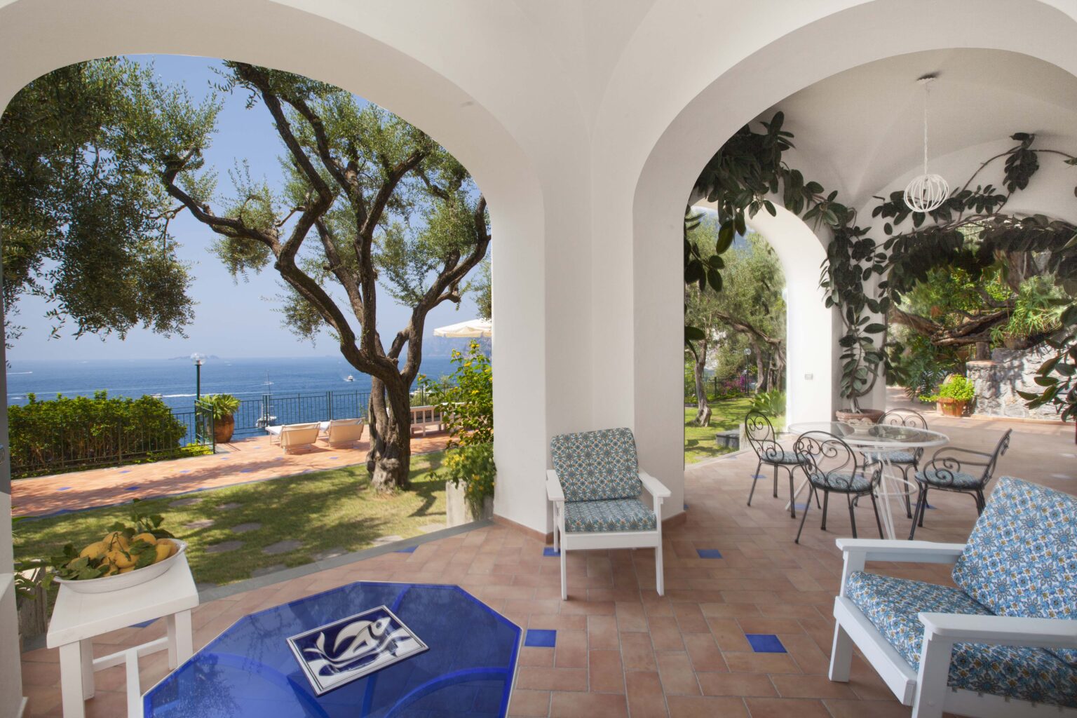 Cozy outdoor seating area with blue and white chairs, overlooking a scenic view of the ocean and lush greenery. The architectural design features arched openings and decorative tiles, creating a tranquil atmosphere perfect for relaxation. | Only on SopranoVillas