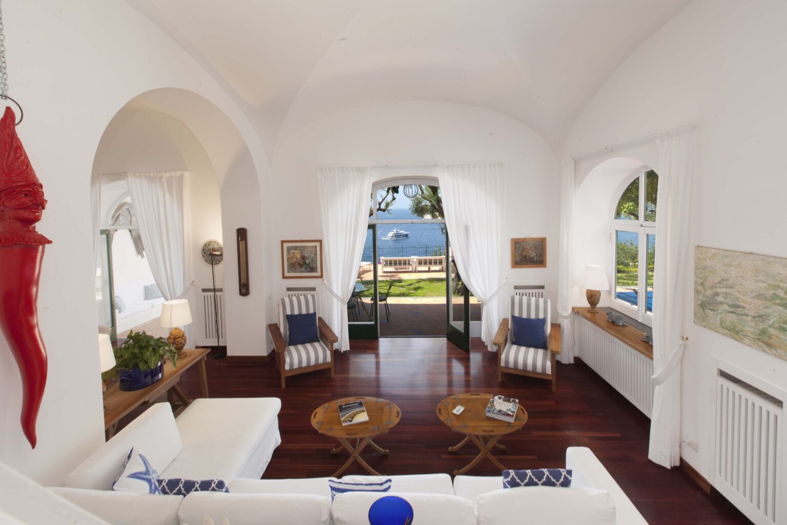 Bright and airy living room featuring white walls and furniture, accented by blue and natural tones. The space includes comfortable seating, wooden coffee tables, and large windows with views of a scenic outdoor area. Ideal for coastal or beach house decor inspiration. | Only on SopranoVillas