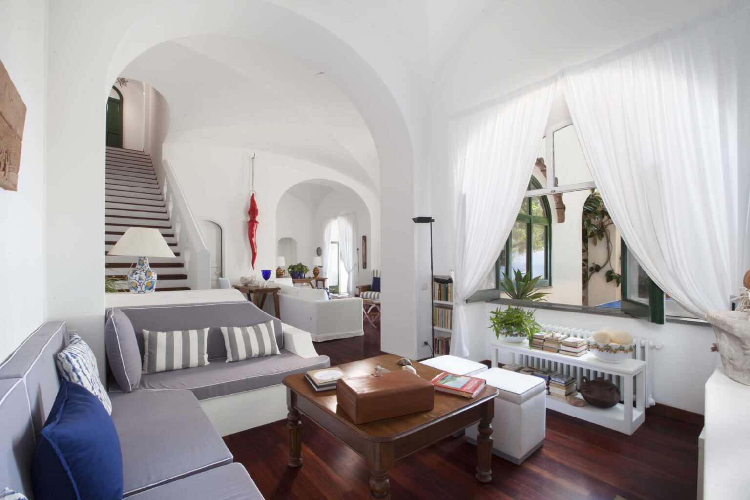 Bright and airy living room featuring a comfortable sofa with decorative pillows, a wooden coffee table, and large windows draped with white curtains. The space includes a staircase in the background and indoor plants, creating a welcoming atmosphere. Ideal for showcasing modern interior design. | Only on SopranoVillas