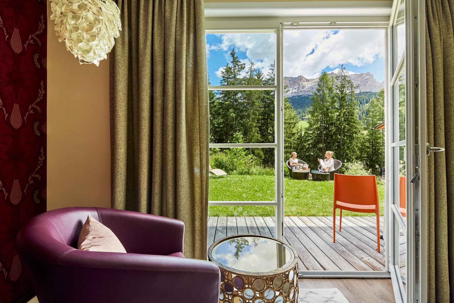 Cozy hotel room featuring a purple armchair and a small table, with large glass doors opening to a scenic outdoor area. A couple enjoys the view of mountains and greenery from the patio, creating a tranquil atmosphere perfect for relaxation. | Only on SopranoVillas