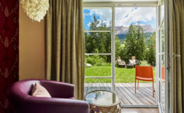 Cozy hotel room featuring a purple armchair and a small table, with large glass doors opening to a scenic outdoor area. A couple enjoys the view of mountains and greenery from the patio, creating a tranquil atmosphere perfect for relaxation.