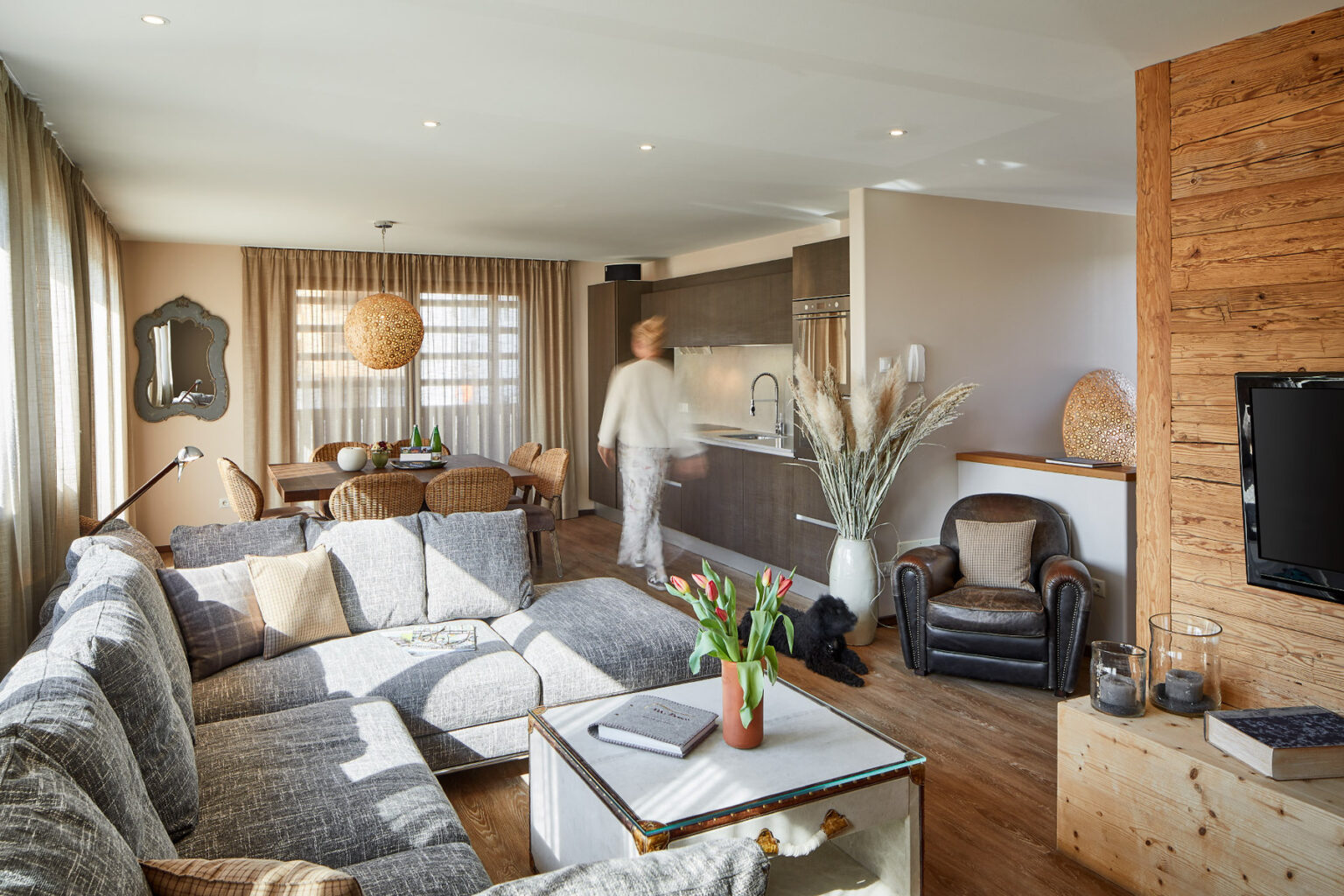 Modern living room featuring a gray sectional sofa, a wooden dining table with wicker chairs, and a stylish kitchen. Natural light floods the space, highlighting decorative elements like a large pendant light, potted plants, and a cozy armchair. A person is seen moving through the room, adding a sense of life to the contemporary interior design. | Only on SopranoVillas