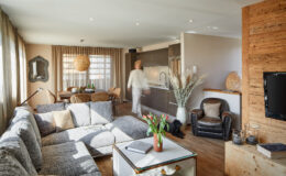Modern living room featuring a gray sectional sofa, a wooden dining table with wicker chairs, and a stylish kitchen. Natural light floods the space, highlighting decorative elements like a large pendant light, potted plants, and a cozy armchair. A person is seen moving through the room, adding a sense of life to the contemporary interior design.