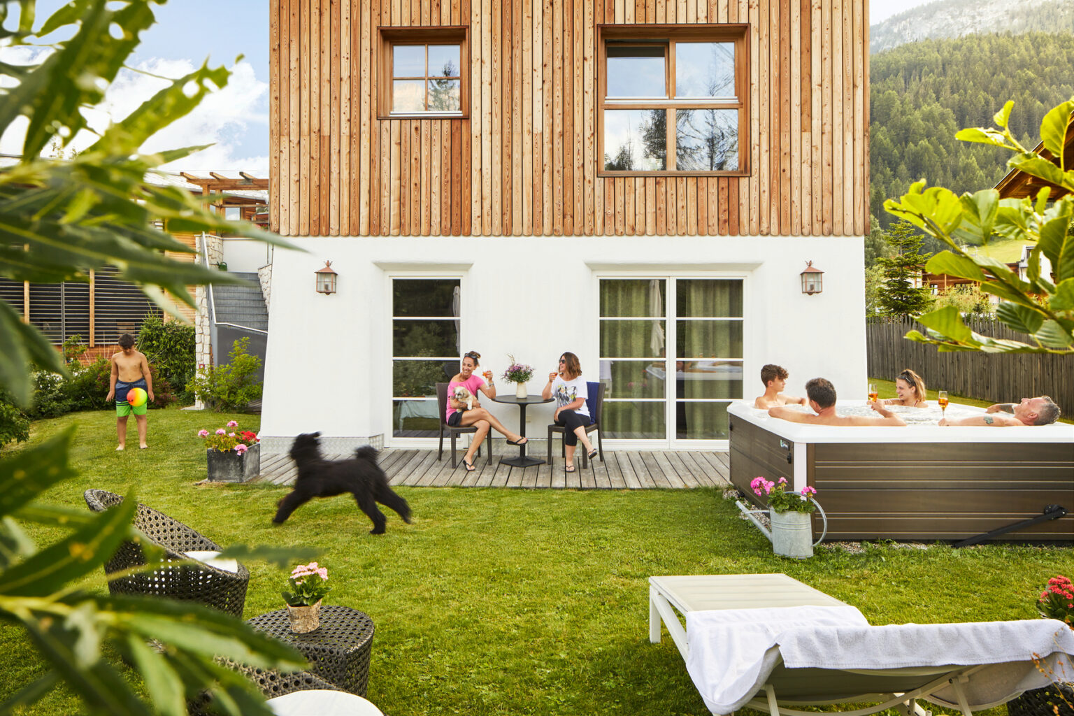 A modern wooden house with a spacious backyard featuring a hot tub, where a group of people relax and socialize. A child is playing with a dog on the lawn, while others sit at a table enjoying drinks. Lush greenery surrounds the outdoor space, creating a serene atmosphere. | Only on SopranoVillas