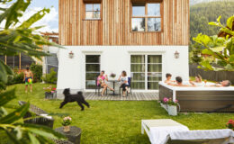 A modern wooden house with a spacious backyard featuring a hot tub, where a group of people relax and socialize. A child is playing with a dog on the lawn, while others sit at a table enjoying drinks. Lush greenery surrounds the outdoor space, creating a serene atmosphere.