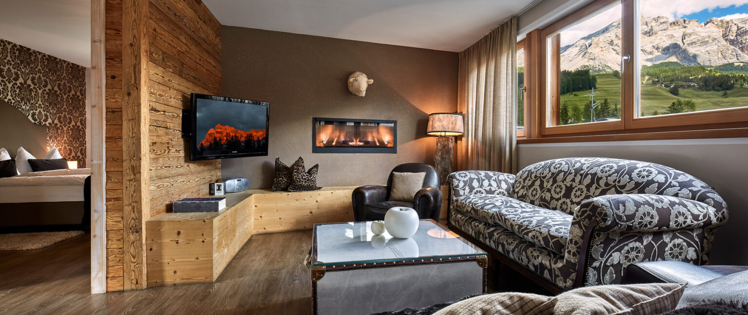 Cozy living room featuring a modern design with wooden accents, a wall-mounted TV, a stylish sofa, and a coffee table. Large windows offer a scenic view of the mountains, enhancing the inviting atmosphere of the space. | Only on SopranoVillas