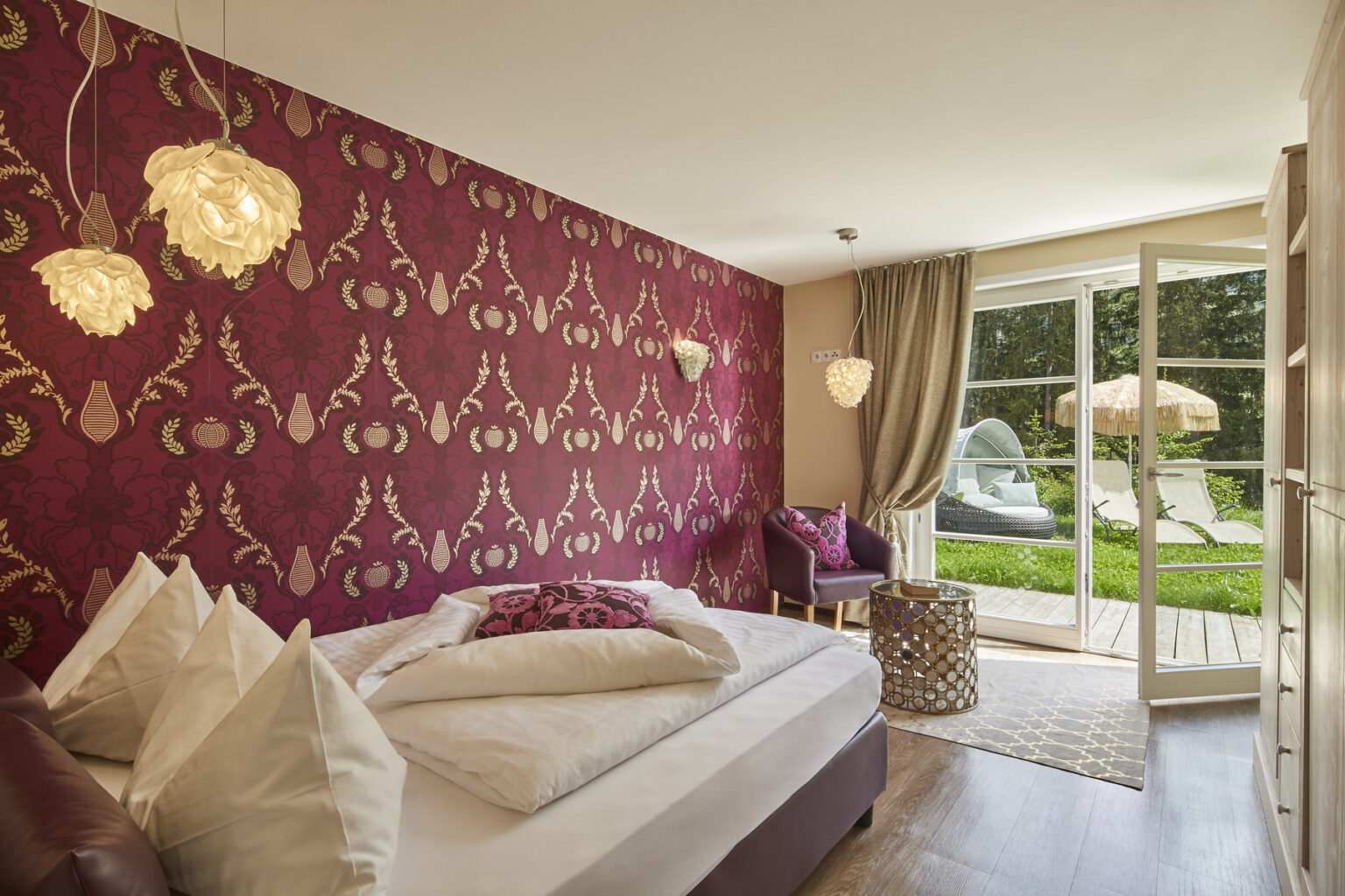 Cozy bedroom featuring a burgundy patterned accent wall, plush bedding with decorative pillows, stylish lighting fixtures, and a view of a green outdoor area with lounge chairs. The room includes a small seating area and modern decor elements, creating a welcoming atmosphere. | Only on SopranoVillas