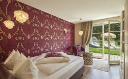 Cozy bedroom featuring a burgundy patterned accent wall, plush bedding with decorative pillows, stylish lighting fixtures, and a view of a green outdoor area with lounge chairs. The room includes a small seating area and modern decor elements, creating a welcoming atmosphere.