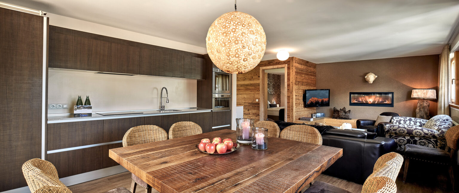 Modern dining area featuring a rustic wooden table with apples and candles, surrounded by woven chairs. A stylish pendant light hangs above, with a contemporary kitchen visible in the background, showcasing dark cabinetry and a sleek design. Cozy living space with a television and wood accents creates a warm atmosphere. | Only on SopranoVillas