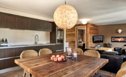 Modern dining area featuring a rustic wooden table with apples and candles, surrounded by woven chairs. A stylish pendant light hangs above, with a contemporary kitchen visible in the background, showcasing dark cabinetry and a sleek design. Cozy living space with a television and wood accents creates a warm atmosphere.