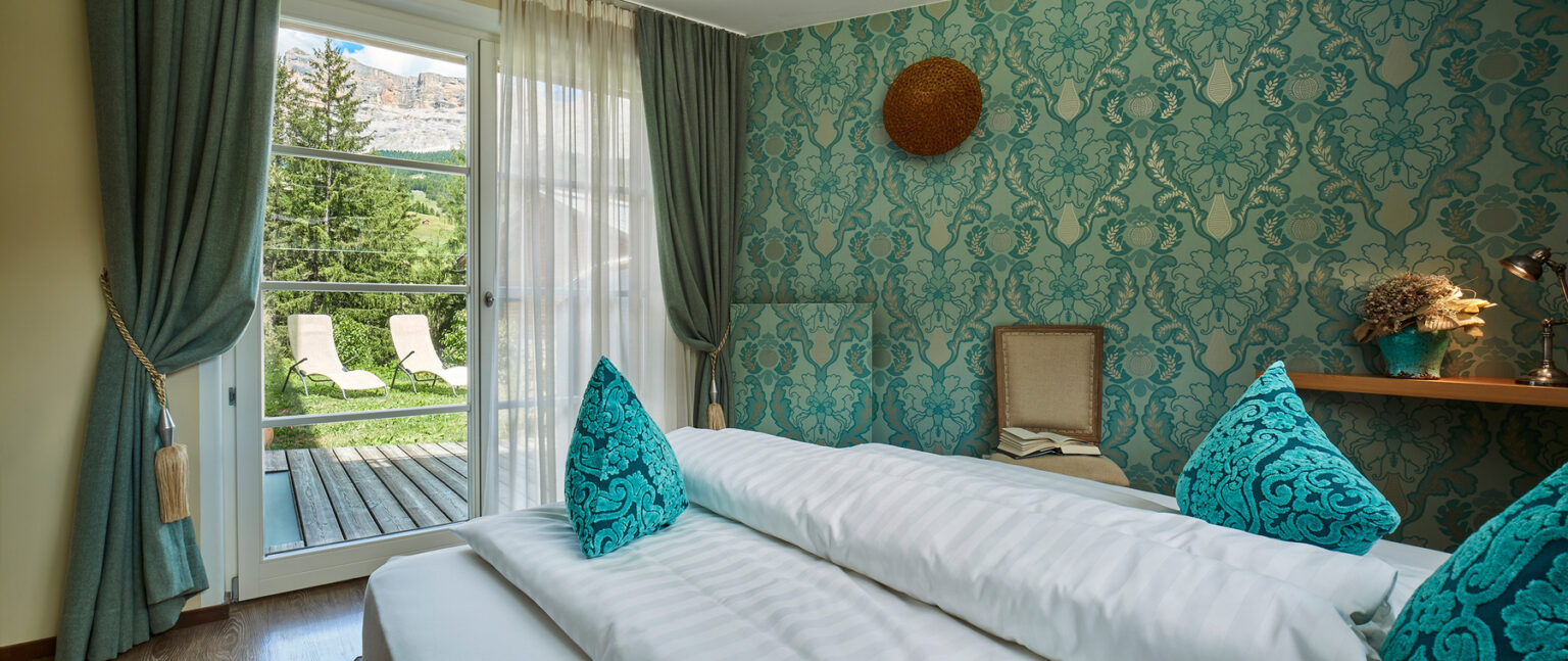 Cozy hotel room featuring a patterned turquoise wall, plush decorative pillows, and a view of outdoor lounge chairs and lush greenery through a large window. The room is designed for relaxation, with elegant curtains and a wooden shelf displaying decorative elements. | Only on SopranoVillas