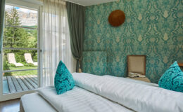 Cozy hotel room featuring a patterned turquoise wall, plush decorative pillows, and a view of outdoor lounge chairs and lush greenery through a large window. The room is designed for relaxation, with elegant curtains and a wooden shelf displaying decorative elements.