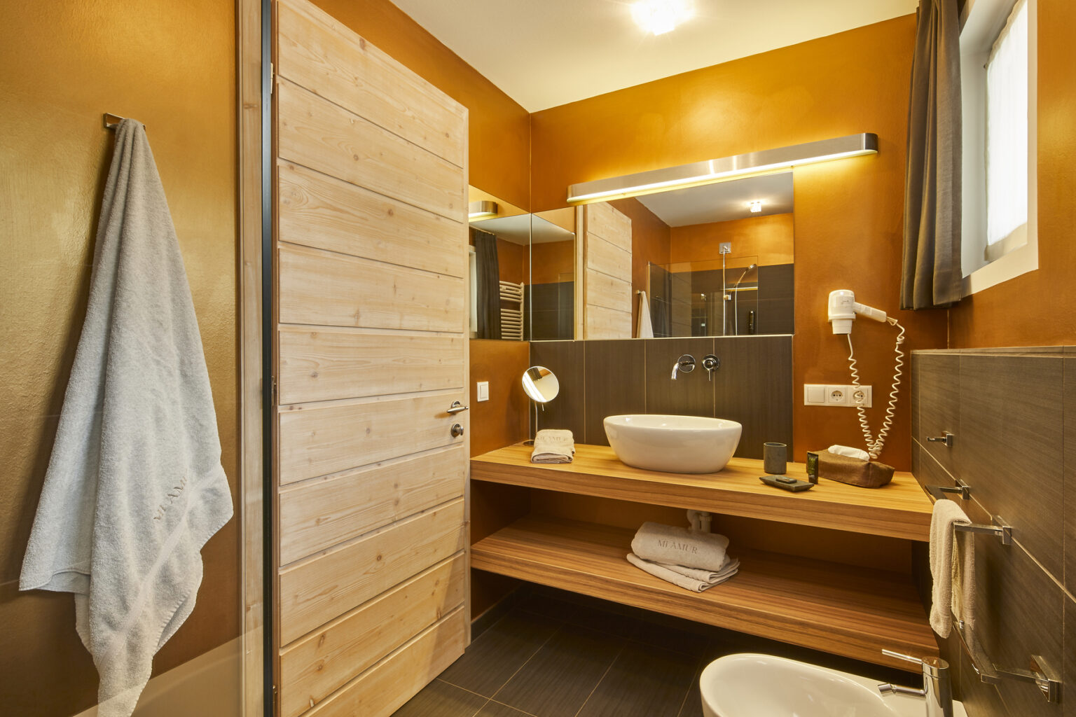 Modern bathroom interior featuring wooden accents, a stylish vessel sink, and warm orange walls, with a towel hanging on the door, providing a cozy and inviting atmosphere. | Only on SopranoVillas