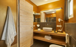 Modern bathroom interior featuring wooden accents, a stylish vessel sink, and warm orange walls, with a towel hanging on the door, providing a cozy and inviting atmosphere.