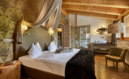 Cozy and elegant hotel room featuring a king-sized bed with plush pillows, warm wooden accents, and stylish decor. Includes a modern bathroom area with glass partitions and a comfortable seating area, ideal for relaxation.
