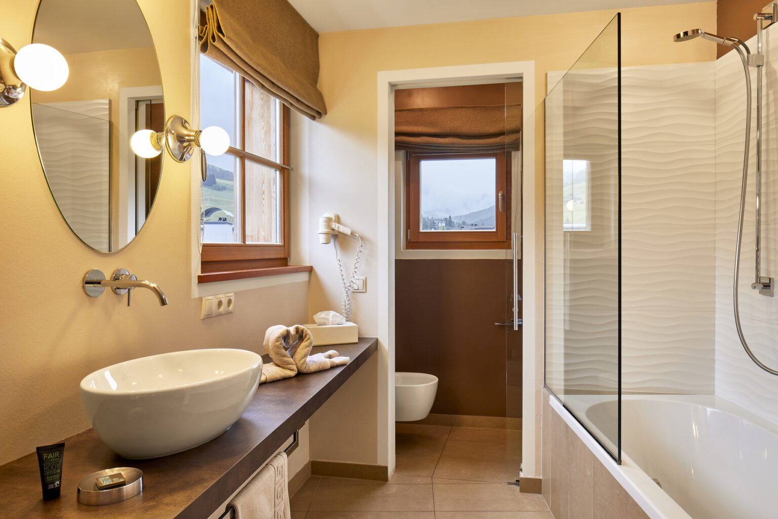 Modern bathroom interior featuring a stylish round sink, wooden countertop, and a combination of a bathtub and shower. Natural light floods the space through a window, complemented by warm beige walls and elegant fixtures. Ideal for showcasing contemporary bathroom design and decor. | Only on SopranoVillas