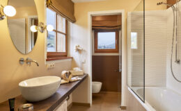 Modern bathroom interior featuring a stylish round sink, wooden countertop, and a combination of a bathtub and shower. Natural light floods the space through a window, complemented by warm beige walls and elegant fixtures. Ideal for showcasing contemporary bathroom design and decor.
