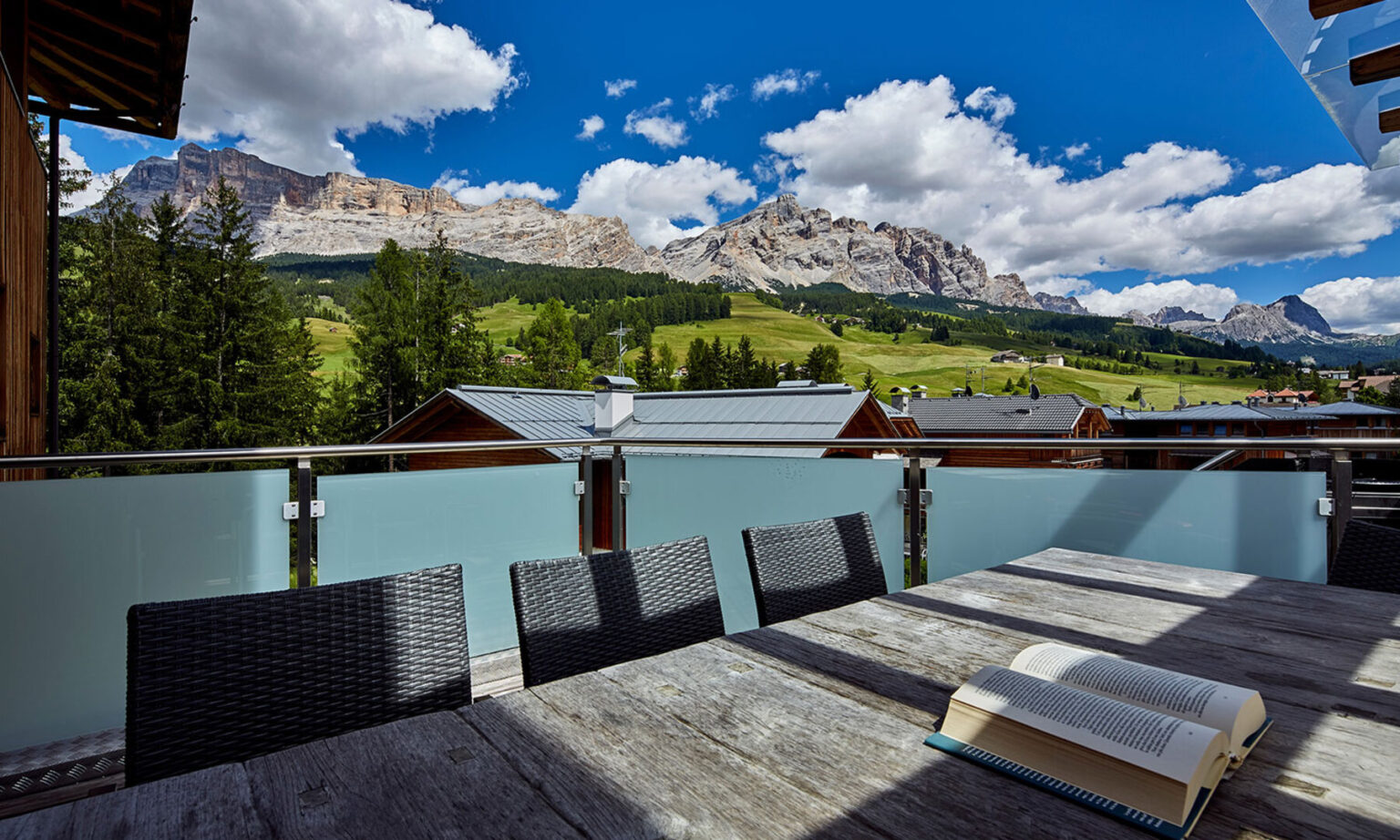Scenic view from a balcony overlooking the Dolomite mountains, featuring a wooden table with an open book and wicker chairs, surrounded by lush greenery and traditional alpine chalets under a bright blue sky with fluffy clouds. | Only on SopranoVillas