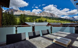 Scenic view from a balcony overlooking the Dolomite mountains, featuring a wooden table with an open book and wicker chairs, surrounded by lush greenery and traditional alpine chalets under a bright blue sky with fluffy clouds.