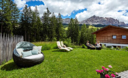 Cozy outdoor seating area with modern lounge chairs and a circular sofa on a lush green lawn, surrounded by tall trees and stunning mountain views under a partly cloudy sky. Ideal for relaxation and enjoying nature.